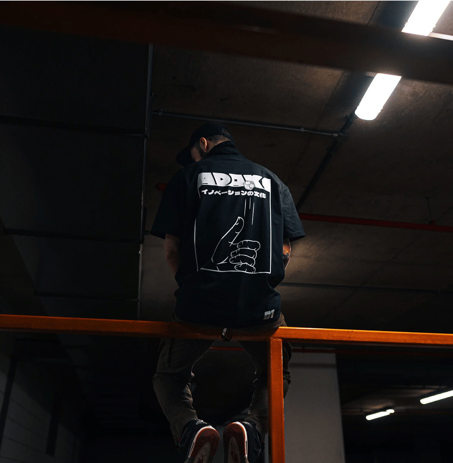 A photograph of the back of a man wearing an Adaki T-shirt, sitting on an orange railing