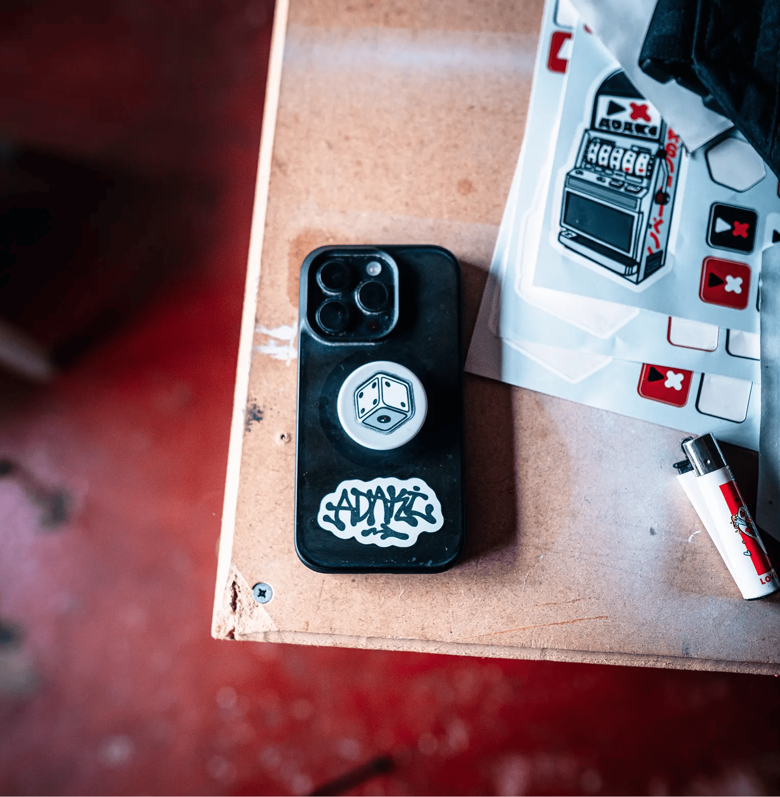 A photograph from a top down view of a phone on a table. The phone is face down, showing Gambler pack stickers of a dice and a graffiti tag of Adaki stuck to the phone case. There is sheets from the gambler pack on the table next to the phone