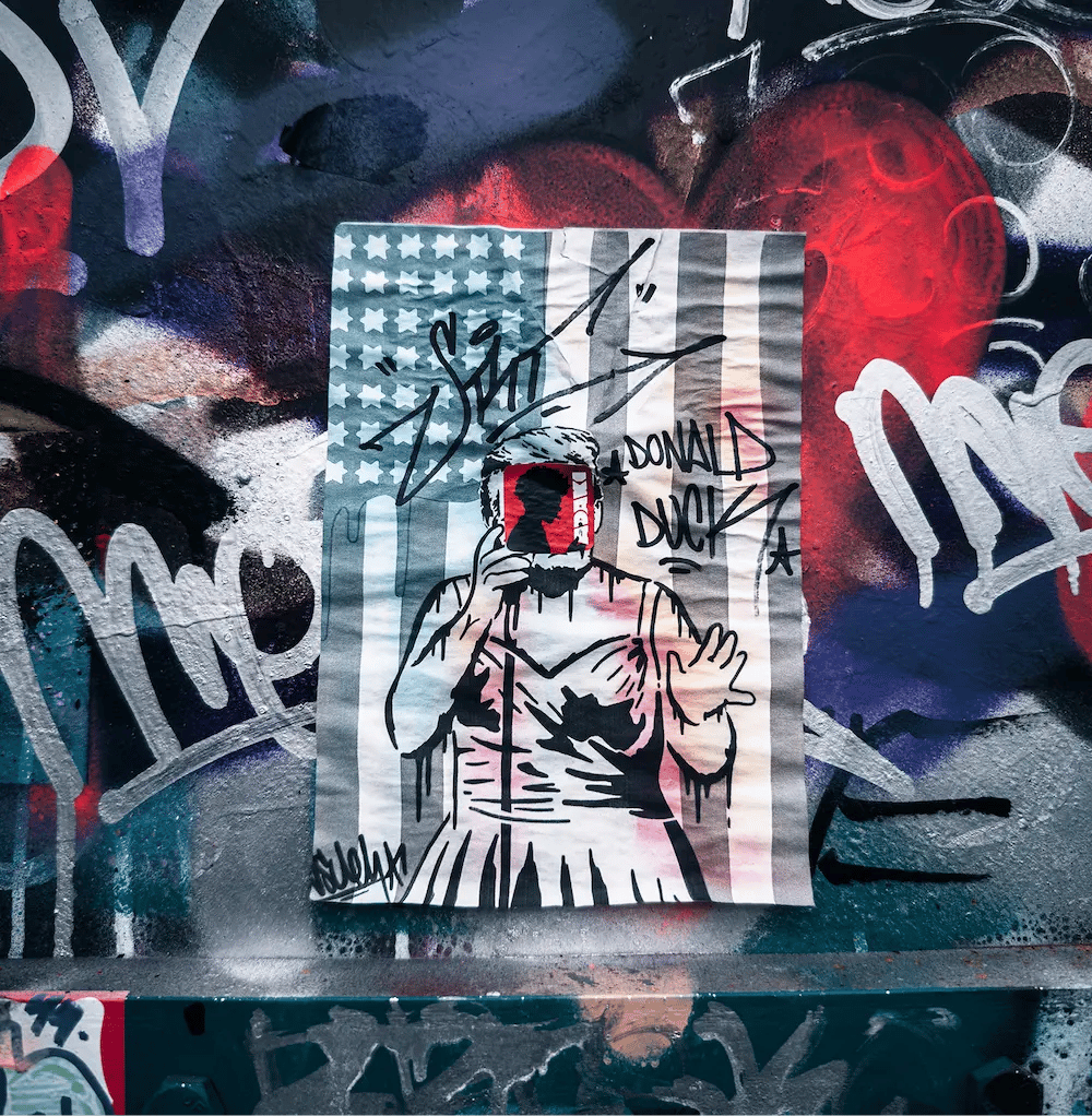A piece of street art on a graffiti-covered wall depicting a figure in a dress holding a microphone against a faded American flag background. The figure’s face is obscured with an Adaki silhouette sticker, and the artwork is defaced with graffiti tags including 'SPIT' and 'DONALD DUCK'. The surrounding wall is layered with colorful graffiti and tags.