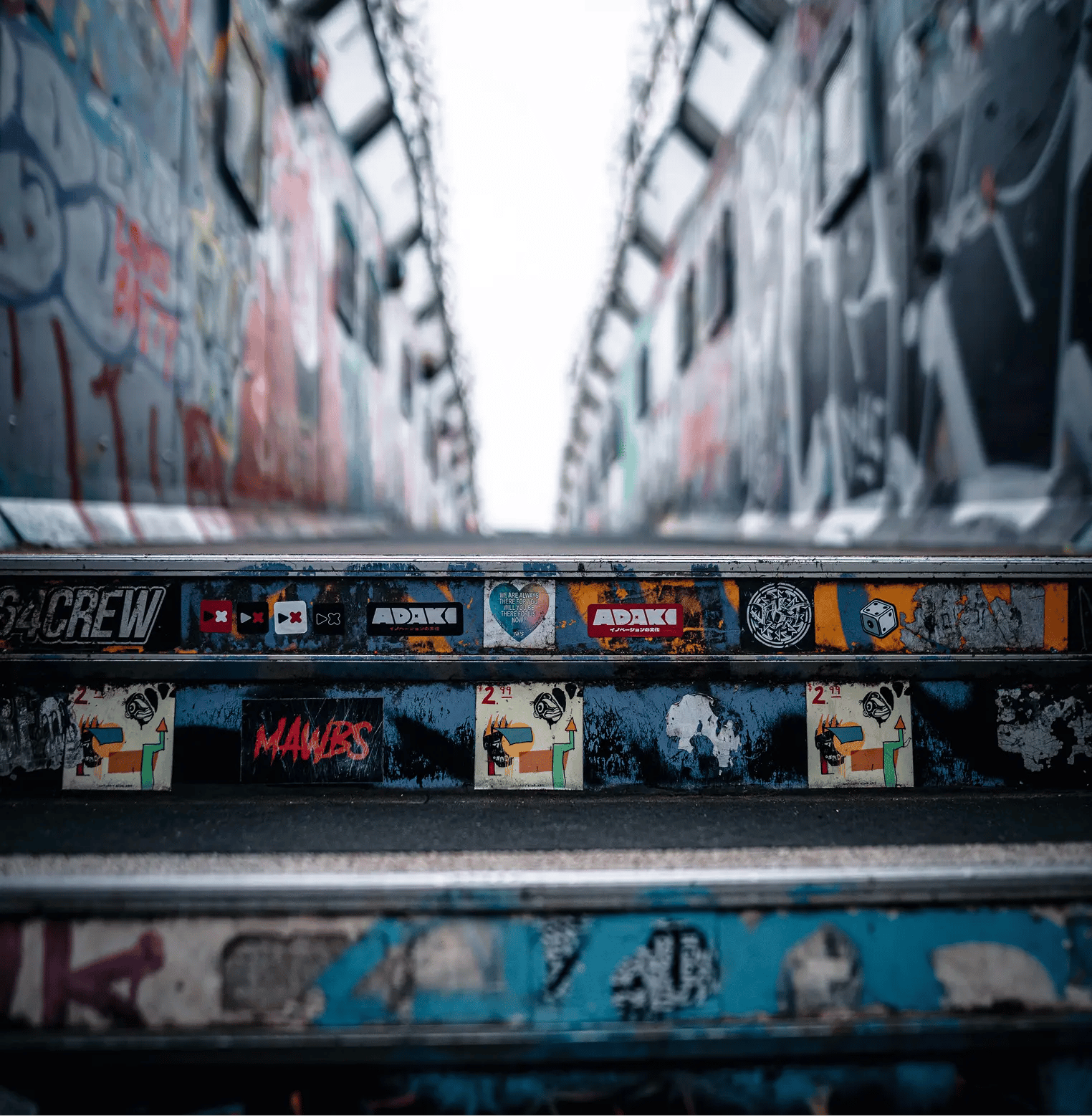 A close-up view of metal stairs covered in stickers and graffiti, with walls of a passageway on either side also covered in graffiti. Prominently visible are multiple 'ADAKI' brand stickers on the stair risers, alongside other designs including hearts, dice, faces, and various tags. The background fades into a bright, overcast sky at the top of the stairway.