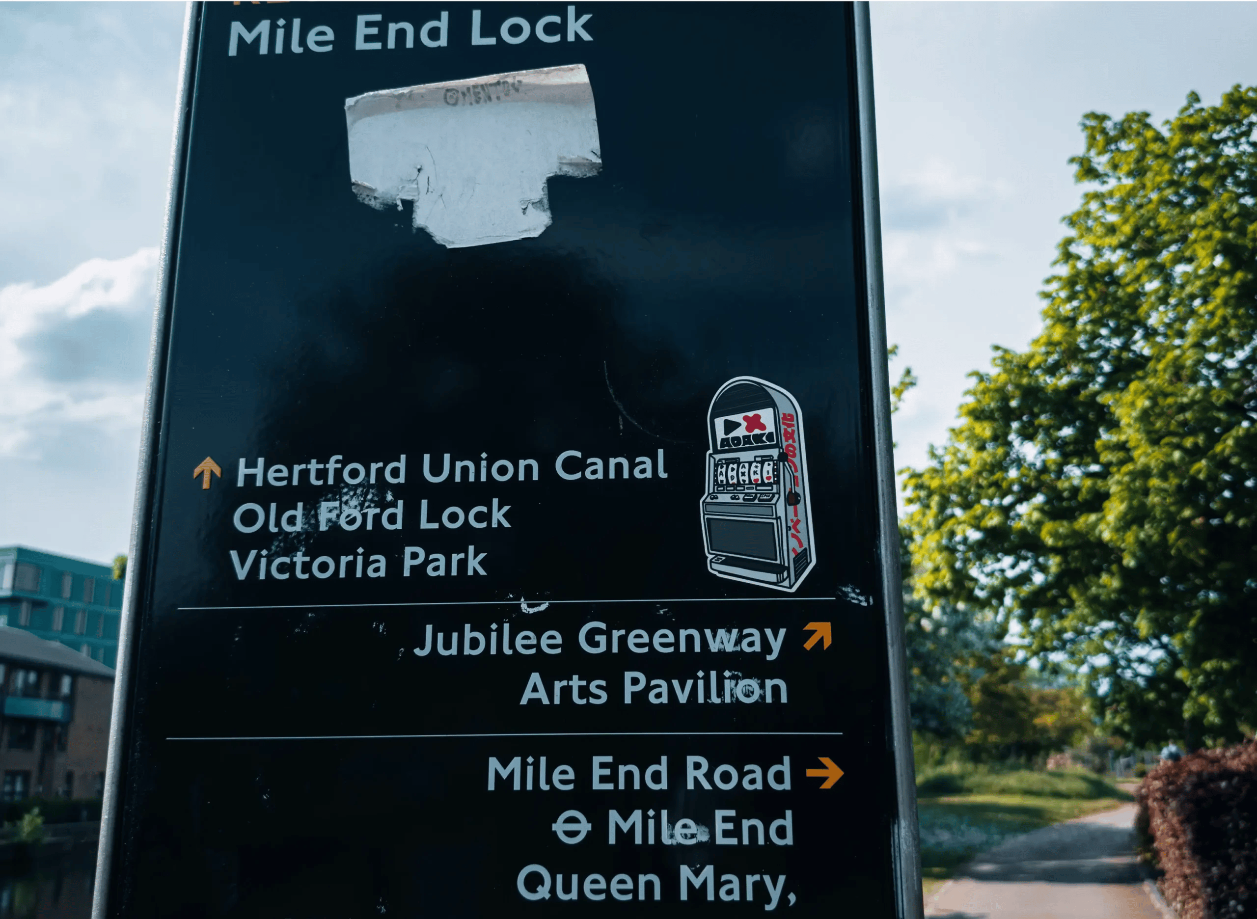 A black directional sign near Mile End Lock with white and orange text pointing to local landmarks like Hertford Union Canal, Victoria Park, and Mile End Road. On the right side of the sign is an 'Adaki' sticker featuring a one-arm bandit slot machine design, with 'Adaki' branding across the top of the machine. Trees and a path are visible in the background under a blue sky.