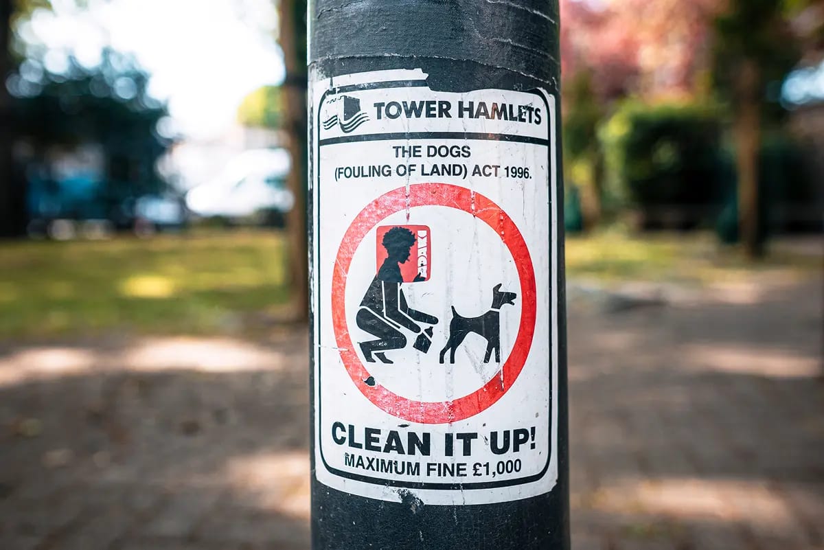 A photograph of a 'clean it up' dog fouling sign in Tower Hamlets London. An Adaki sticker of the silhouette of a character is stuck over the head of the man picking up dog foul on the sign.