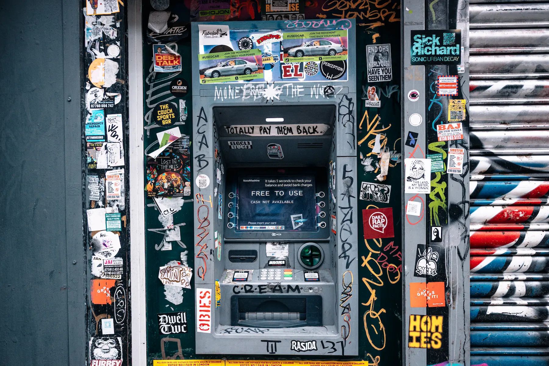 An ATM machine covered in graffiti, stickers, and street art, situated between two heavily stickered walls. The ATM screen displays a message stating 'FREE TO USE - CASH NOT AVAILABLE'. Surrounding the ATM are various colourful stickers, posters, and handwritten tags, contributing to an urban, gritty aesthetic. Adaki gambler pack stickers are visible in multiple places.
